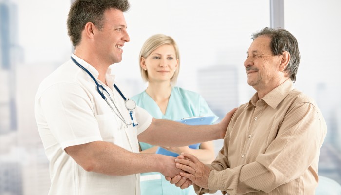 Smiling medical doctor shaking hands with happy senior patient, nurse in background.
